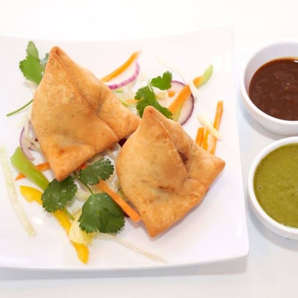 Two golden-brown samosas on a white plate with shredded vegetables and two dipping sauces.